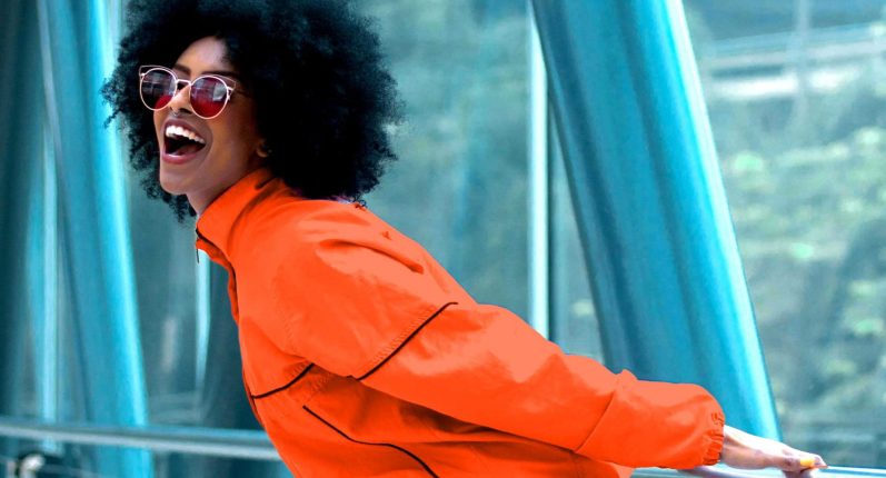 Eine Frau mit Afrofrisur und orange-farbener Jacke und Sonnenbrille, lacht fröhlich.